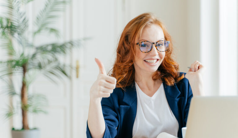 Positive redhead woman keeps thumb raised, demonstrates like gesture, satisfied with good work of colleague, updates software on modern gadget, searches data on website, poses in coworking space